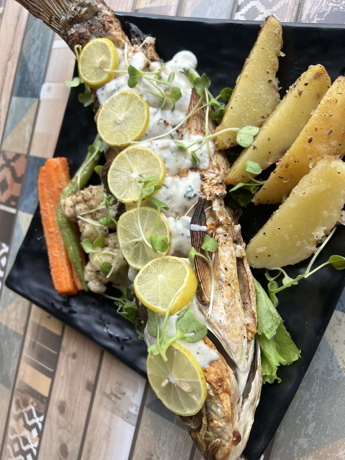A beautifully presented whole grilled fish served on a black platter, draped in a creamy white sauce and topped with fresh lemon wheels and microgreens. Thick, seasoned potato wedges and grilled carrots line the side. — DASH 식단 하루 구성, 뭘 얼마나 먹나